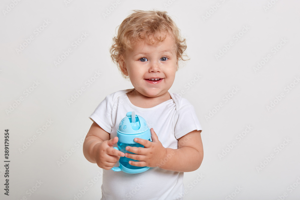 Portrait of cute toddler holding water bottle, kid playing with blue baby cup, adorable curly hair boy being thirsty, looking at camera with toothy smile and curious facial expression. Portrait of cute toddler holding water bottle, kid playing with blue baby cup, adorable curly hair boy being thirsty, looking at camera with toothy smile and curious facial expression.