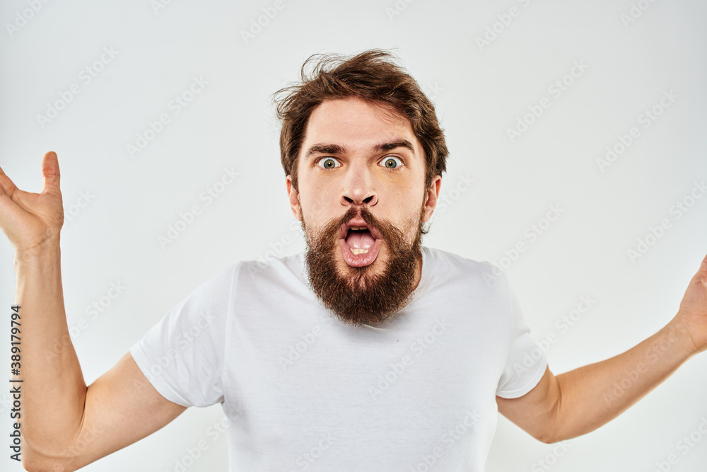 Emotional bearded man in a white t-shirt gestures with his hands light background