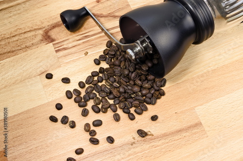 Top-down view of manual coffee grinder laying on wooden table with spilled coffee beans around. Concept of fresh coffee culture.