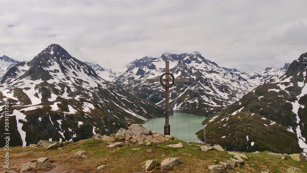 Beautiful panorama view of Silvretta lake, Montafon, Austria and the surrounding snow-covered mountains from peak Bielerspitze with wooden summit cross in front.