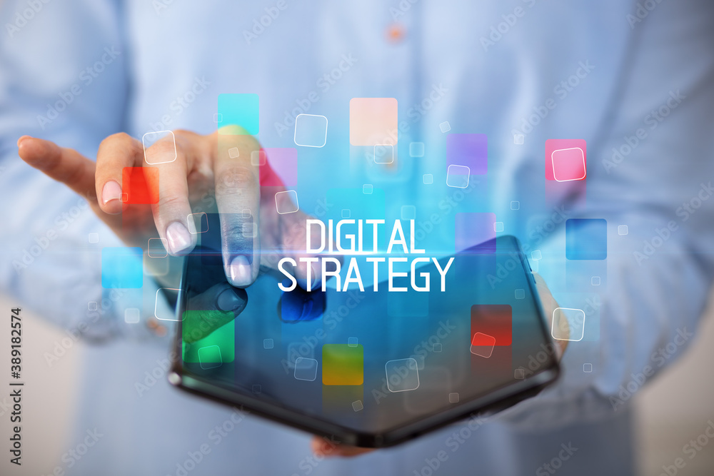 Young man holding a foldable smartphone with DIGITAL STRATEGY inscription, educational concept Young man holding a foldable smartphone with DIGITAL STRATEGY inscription, educational concept