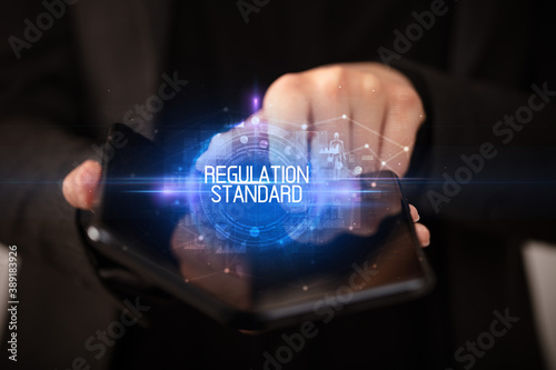 Young man holding a foldable smartphone with REGULATION STANDARD inscription, educational concept