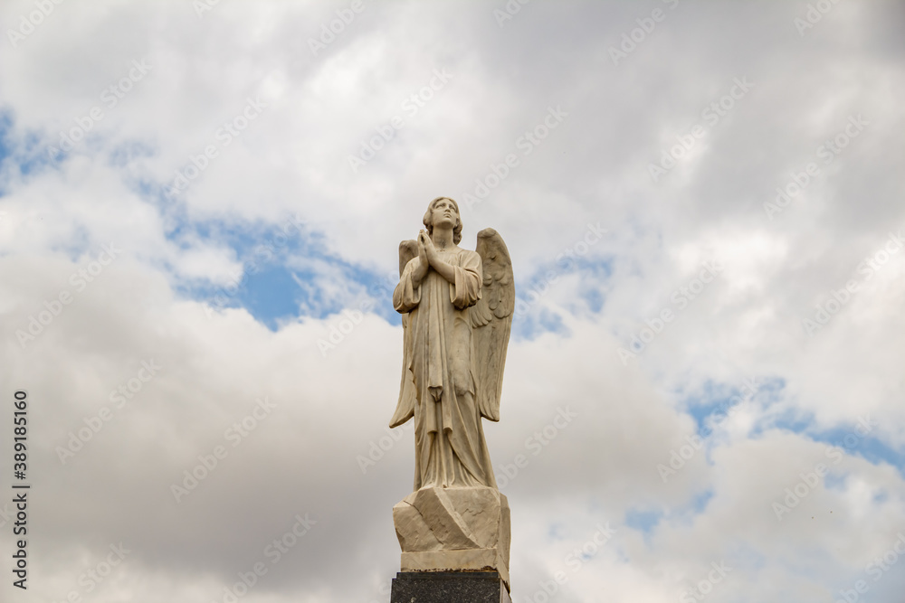 Escultura de anjo em topo de mausoléu dentro de um cemitério com céu nublado ao fundo.