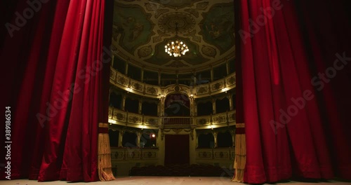 Cinematic shot of empty classic theatre with red velvet curtains opening stage with dramatic lighting before start of show.