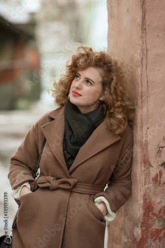 Portrait of a young woman with curly hair leaning against a brick wall. Outside.