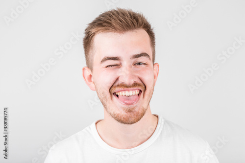 Positive funny male in white shirt, winks an eye and laughs at camera, demonstrates that everything is fine