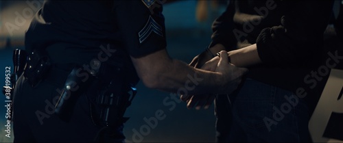CLOSE UP Police officer handcuffs a suspect near police car, African-American Black criminal. Lights flashing in the background. Shot with anamorphic lens photo