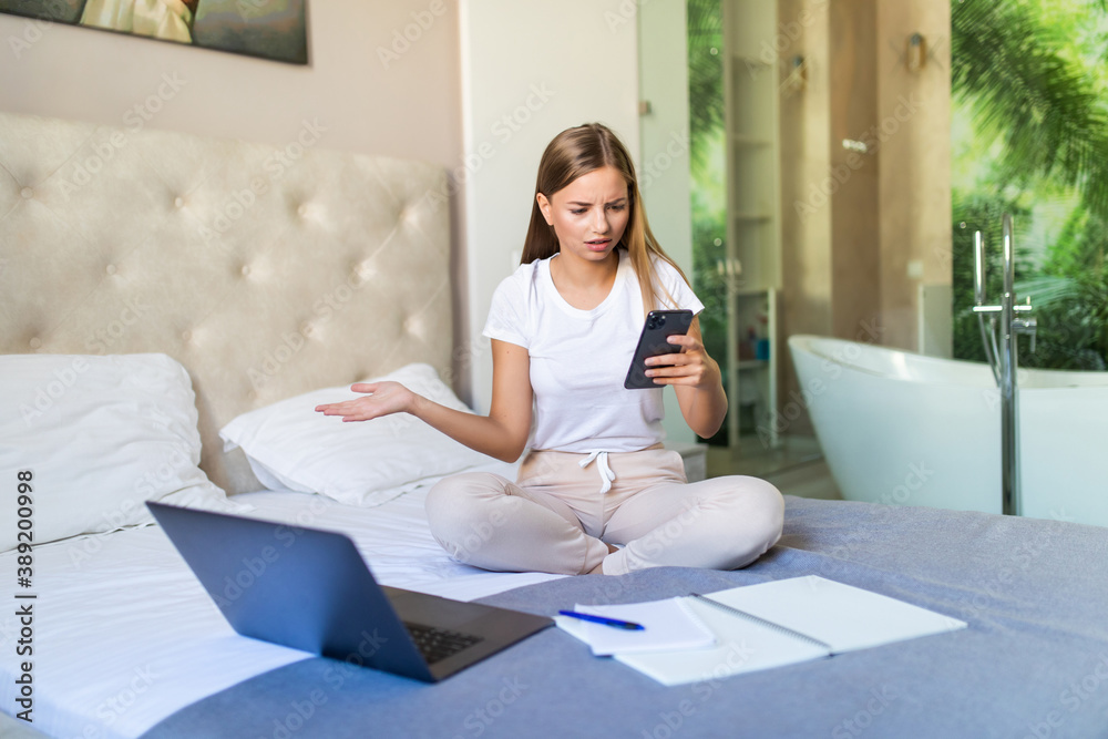 Young pretty woman sitting on bed use mobile phone and laptop while working from home Young pretty woman sitting on bed use mobile phone and laptop while working from home