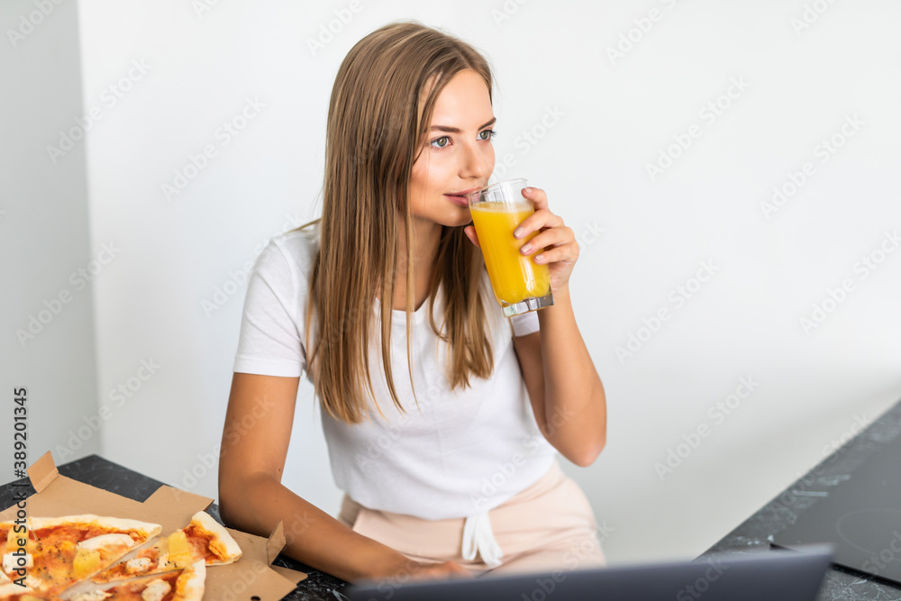 Young woman drink juice and eating pizza and looking at laptop in the kitchen Young woman drink juice and eating pizza and looking at laptop in the kitchen