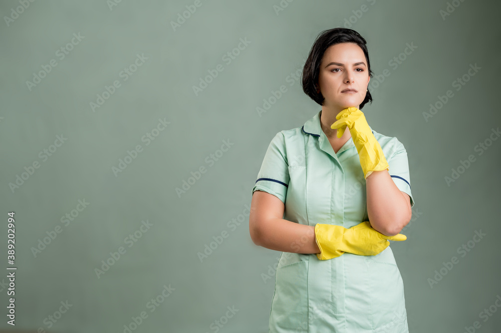Young cleaning woman wearing a green shirt and yellow gloves holding hand under her chin, having doubtful Young cleaning woman wearing a green shirt and yellow gloves holding hand under her chin, having doubtful