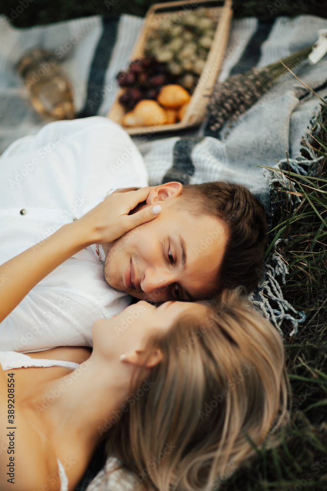 Loving couple lying and cuddling on picnic blanket outdoors. Nearby lies basket with grapes, croissants and white wine.
