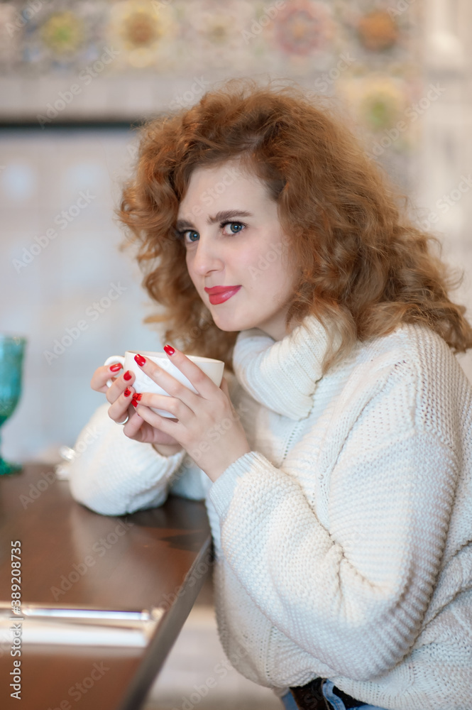 A beautiful girl in a white knitted sweater sits in a cafe with a white cup in her hands. Inside A beautiful girl in a white knitted sweater sits in a cafe with a white cup in her hands. Inside