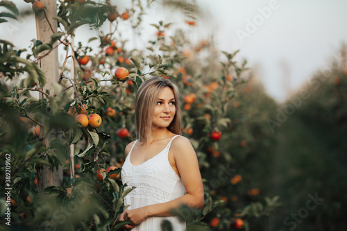 Beautiful young blonde woman in white dress posing near apples trees in the garden.