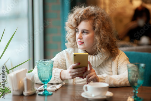 A beautiful girl is sitting in a cafe with a cup of tea and is typing text in the mobile. Inside