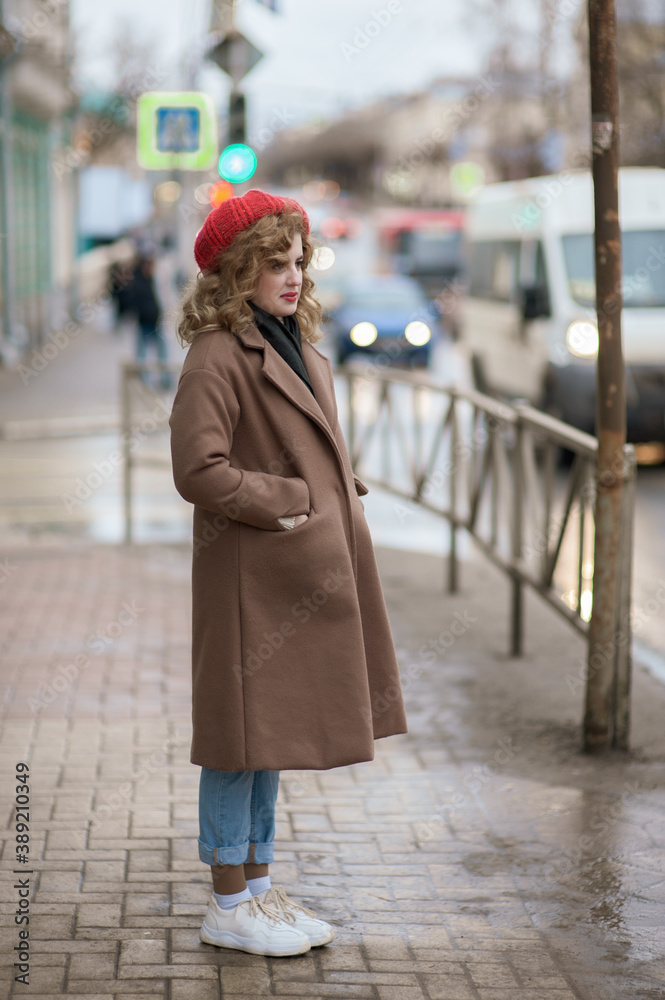 Cute girl in a red hat and brown coat is waiting near the road