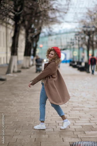 Beautiful brunette young woman in a red hat, jeans, and a coat happily walks along the street