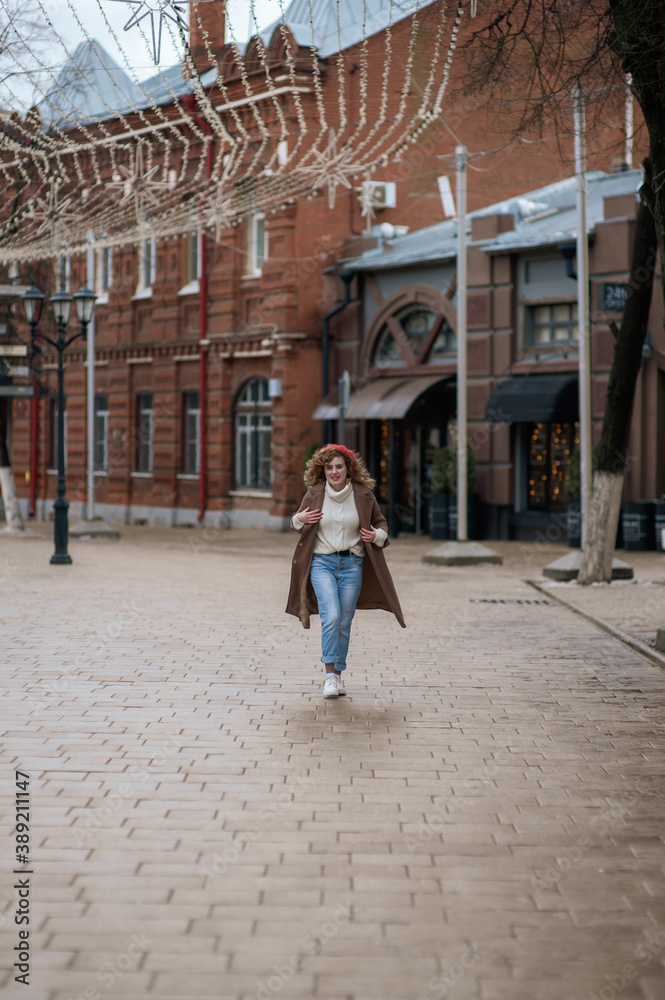 Beautiful brunette young woman in a red hat, jeans, and a coat happily walks along the street