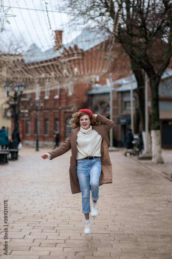 Beautiful brunette young woman in a red hat, jeans, and a coat happily runs down the street