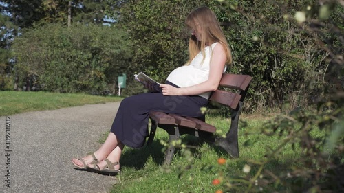 A pregnant woman is sitting on the bench in the park and reading a book. Pregnant woman in the summer park reading.