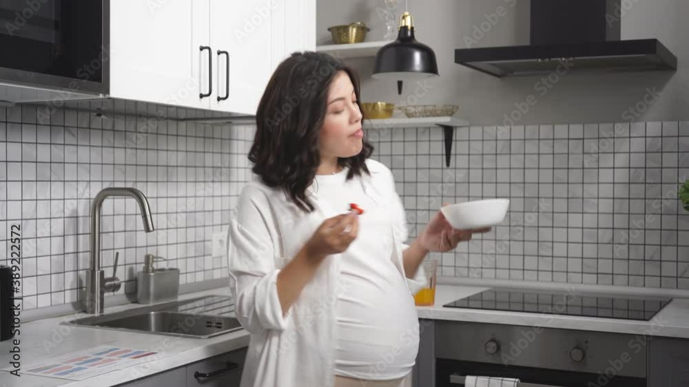 Cheerful pregnant woman happily eat berries in the kithcen, enjoy, hold plate in hands, looks up. Cheerful pregnant woman happily eat berries in the kithcen, enjoy, hold plate in hands, looks up.