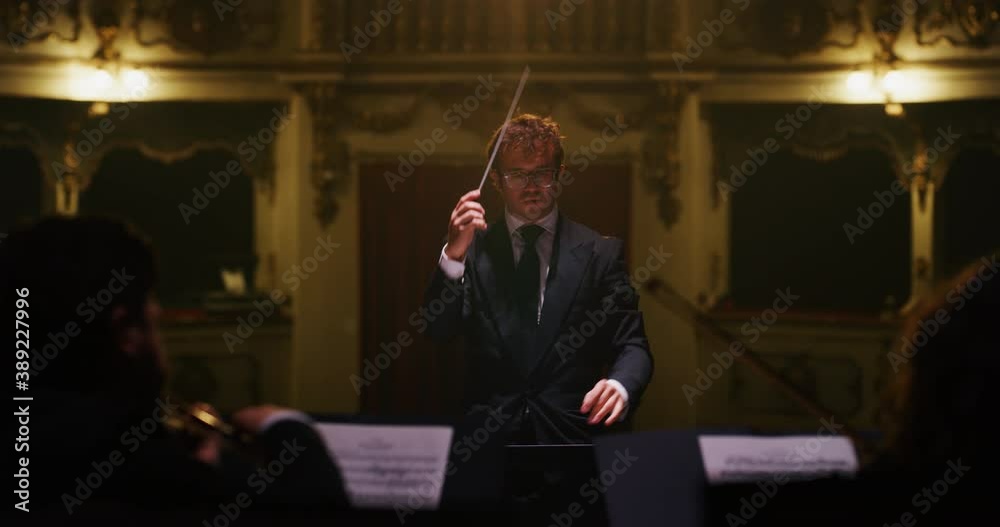 Cinematic shot of conductor directing symphony orchestra with performers playing violins, cello and trumpet on classic theatre with curtain stage during music concert with dramatic lights.