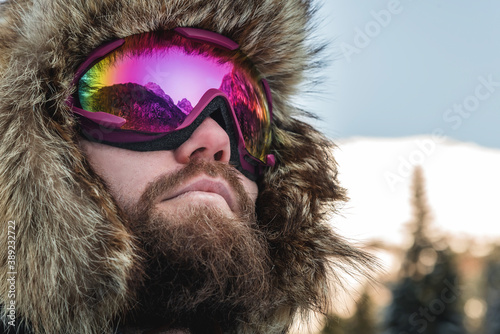 An adult brutal man with a beard in ski goggles and a hat stands in the mountains in winter. Active recreation concept
