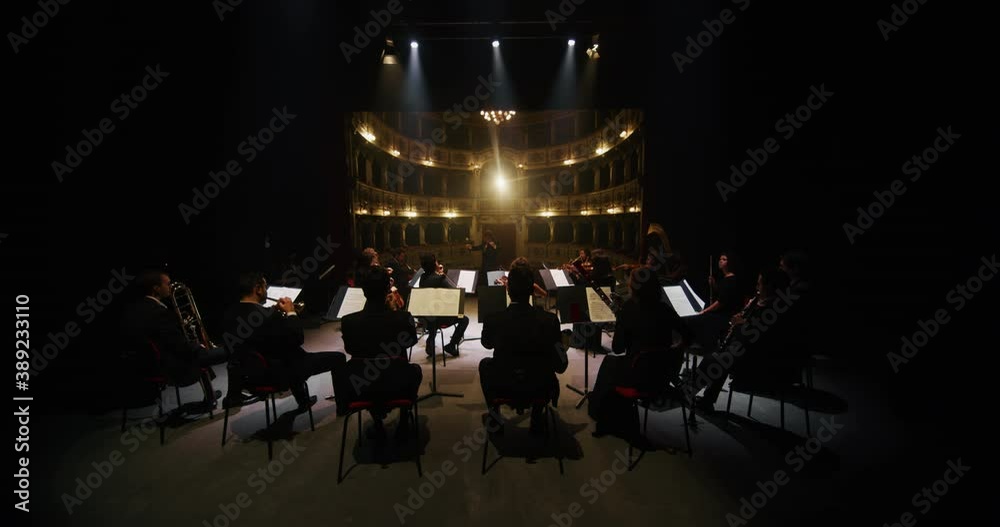 Cinematic shot of conductor directing symphony orchestra with performers playing violins, cello and trumpet on classic theatre with curtain stage during music concert with dramatic lights.