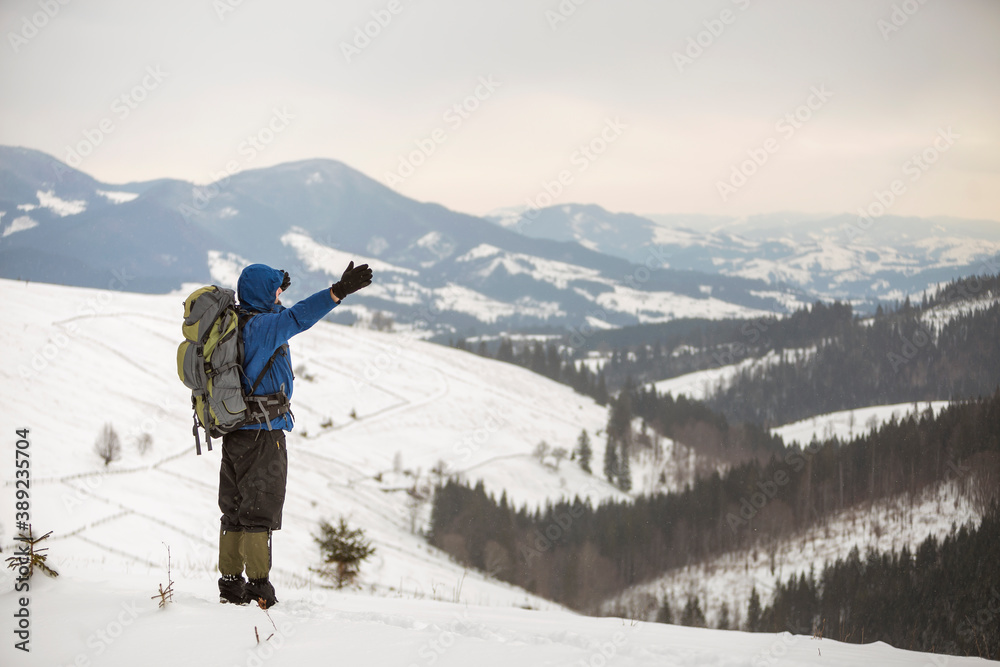 Back view of tourist hiker in warm clothing with backpack standing with raised arms on mountain clearing on copy space background of woody mountain ridge and cloudy sky.