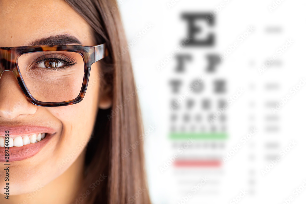 Cloese up of young woman wearing eyeglasses. Chart for eye sight testing in background. Portrait of a female optometrist standing beside an eye test Cloese up of young woman wearing eyeglasses. Chart for eye sight testing in background. Portrait of a female optometrist standing beside an eye test
