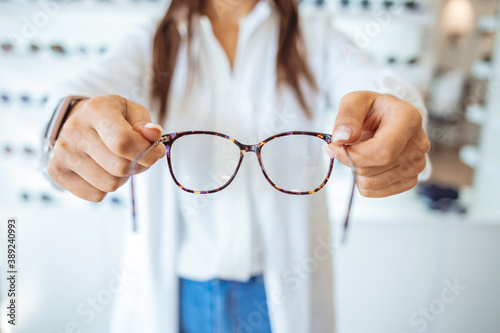 Optician showing and suggesting eyeglasses in optical shop. Cheerful female ophthalmologist is working with patient. Optician giving new glasses to customer for testing and trying.