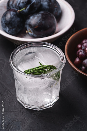 Fresh ice cold carbonated water in glass with rosemary leaf near to wooden bowls with grape and plum fruits, dark stone background, angle view