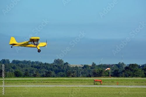 Small  yellow Karatoo Crop checker propeller airplane  low above the runway near a windsock coming in for landing