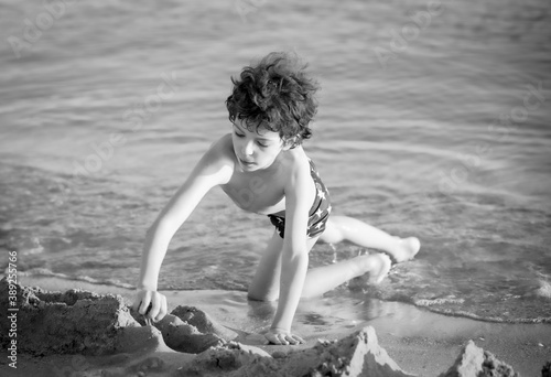 Cute curly male kid playing game at the beach.Little boy play with sand on summer beach.Funny games during summer vacation. Child having fun at coast. Leisure, games and vacation.Black and white photo