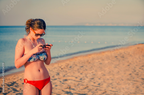 Pretty teenager girl brunette using a smart phone standing on the beach with the sea and horizon in the background. Copy space