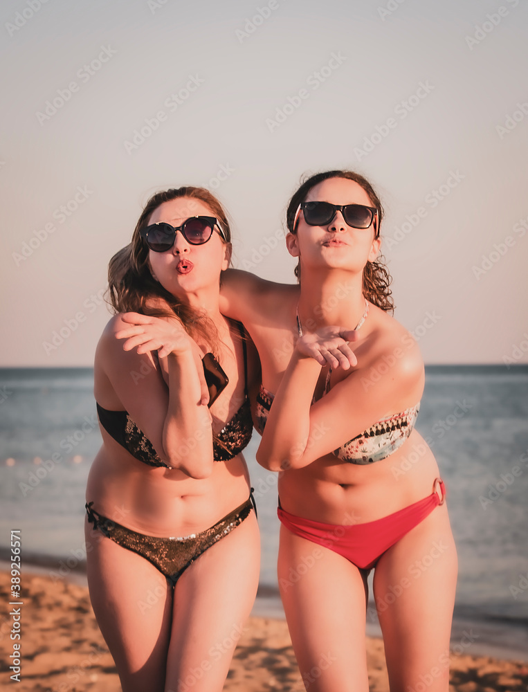 Mom and teenage daughter, looking at the camera and blowing you a kiss, have a slim body, wear bikini sunglasses, posing in front of a tropical beach. The concept of family happiness Mom and teenage daughter, looking at the camera and blowing you a kiss, have a slim body, wear bikini sunglasses, posing in front of a tropical beach. The concept of family happiness