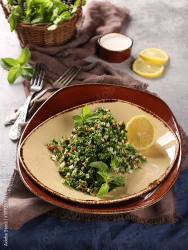 Oriental cuisine. The tabouleh salad. Salad with fresh herbs, bulgur and tomatoes, lemon and olive oil in a light beige plate on a gray table. Fresh mint, sauce, matsoni. Restaurant. Background image