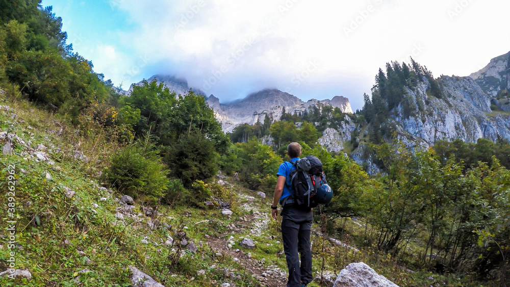 A man hiking in lush green Alpine region in Austria. The sky is blue, with one cloud. High peaks in front of him. Mountains slopes overgrown with bushes and grass. Summer in Alps. Exploring the nature