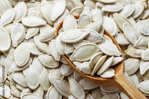 Full frame background texture of fresh pumpkin seeds with a rustic wooden spoon arranged diagonally on the surface.