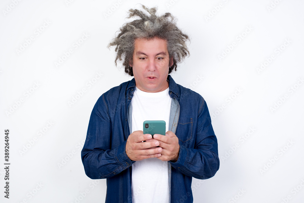Handsome mature caucasian man with afro gray hair standing over white background looks with bugged eyes, holds modern smart phone, receives unexpected message from friend, reads reminder. Handsome mature caucasian man with afro gray hair standing over white background looks with bugged eyes, holds modern smart phone, receives unexpected message from friend, reads reminder.