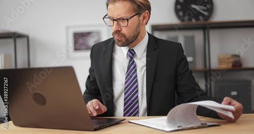 Confused bearded an in business suit and eyglasses rechecking all financial reports while sitting at modern office. Mature male using modern laptop for work. photo