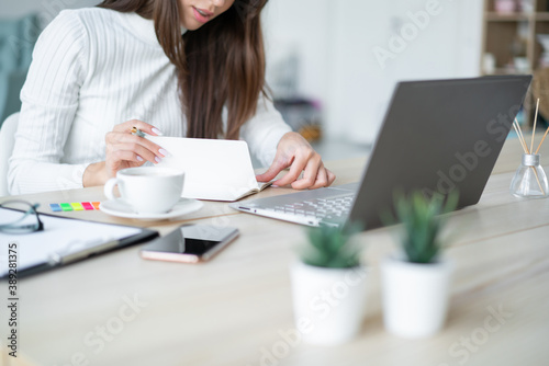 young woman sits desk in front of aptop and turns page of her workbook.