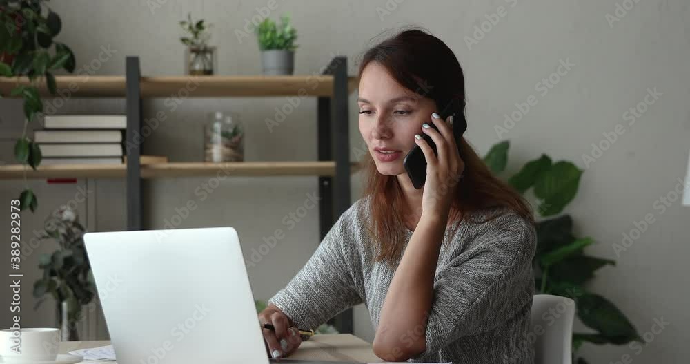 Happy young 30s red-haired businesswoman working on computer, holding mobile phone call conversation with partner colleagues or client, discussing project details or giving professional consultation. Happy young 30s red-haired businesswoman working on computer, holding mobile phone call conversation with partner colleagues or client, discussing project details or giving professional consultation.