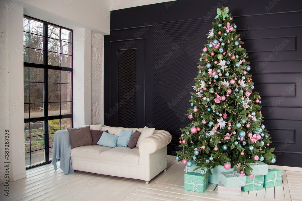 new year's large green Christmas tree decorated with balloons and a garland with gifts and a white sofa with colorful pillows in the loft interior on a dark background with a window.
