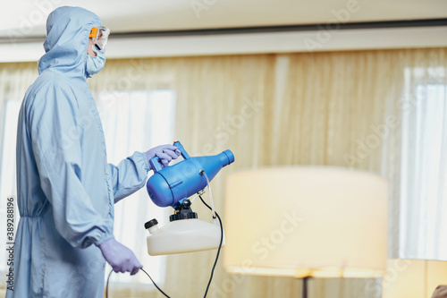 Man in blue protective suit holding disinfectant and standing in the room