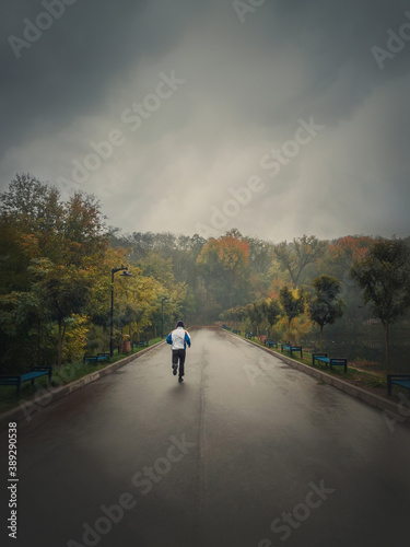 Single man running alone on an alley in the autumn park. Practicing sport outdoors in a rainy and misty day. Gloomy fall season mood. Healthy lifestyle workout jogging activity.