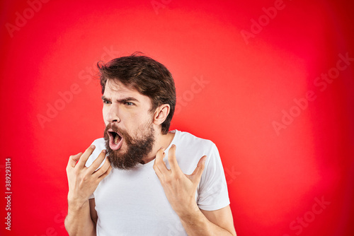 Emotional bearded man gestures with his hands in a white T-shirt aggression discontent red background