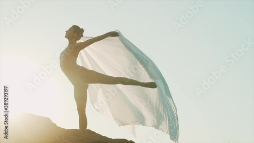 Young beautiful ballerina dancing ballet on clean sky background with huge silk fabric fluttering in wind. Concept of tenderness, lightness, art and talent in nature