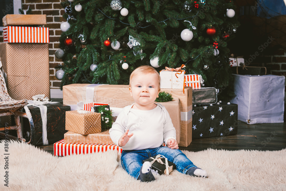 Naklejka premium theme winter and Christmas holidays. Child boy Caucasian blond 1 year old sitting home floor near Christmas tree with New Year decor on shaggy carpet skin receives gifts, opens gift boxes in evening