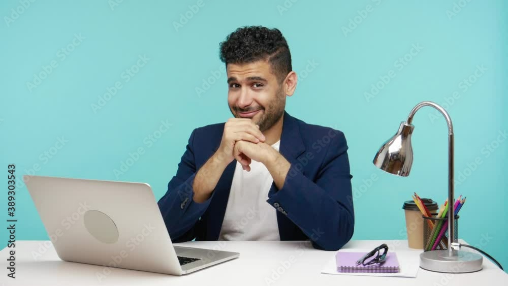 Cunning office worker showing money gesture with fingers sitting at table in his office and looking at camera, teaching how to make money. Indoor studio shot isolated on blue background
