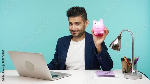 Smiling positive businessman showing piggy bank and thumbs up sitting on workplace in office, satisfied with investments to future. Indoor studio shot isolated on blue background photo